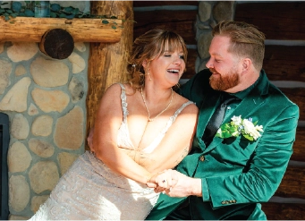 Home A bride in an embellished wedding dress and a groom in a vibrant emerald green suit share a candid laugh while sitting together in front of a rustic stone and log cabin fireplace.