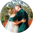 Home A circular crop of a bride in a white gown and a groom in an emerald green velvet suit sharing their first kiss, with snow-streaked mountains and evergreen trees in the background.