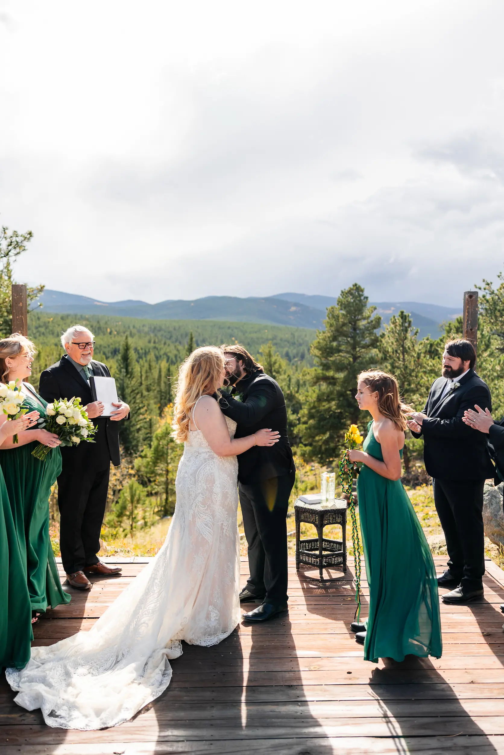 About A bride and groom sharing a first kiss during an outdoor mountain wedding ceremony, surrounded by their bridal party in green dresses.