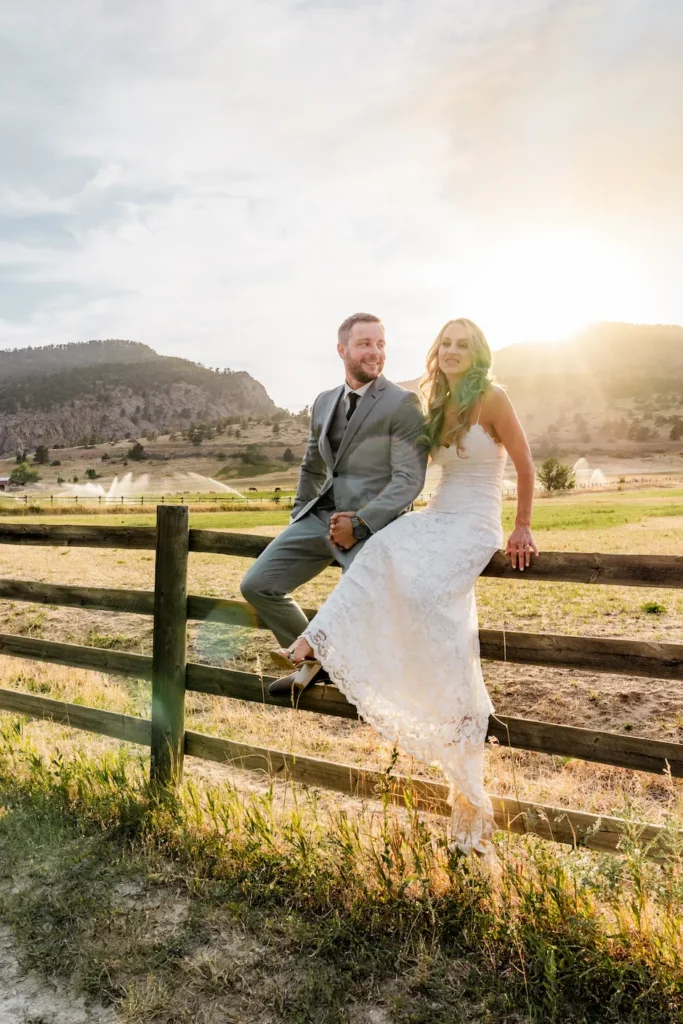 Blog A bride in a white lace dress and a groom in a light grey suit sitting together on a rustic wooden fence, smiling in a sunlit mountain valley.