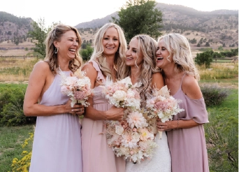 Home A bride laughing and celebrating outdoors with her three bridesmaids in shades of pink and mauve dresses, holding lush floral bouquets with hills in the background.