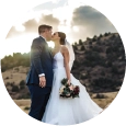 Home A circular crop of a bride in a white wedding gown holding a dark floral bouquet and a groom in a navy suit kissing at sunset, with a golden light flare and rolling hills in the background.