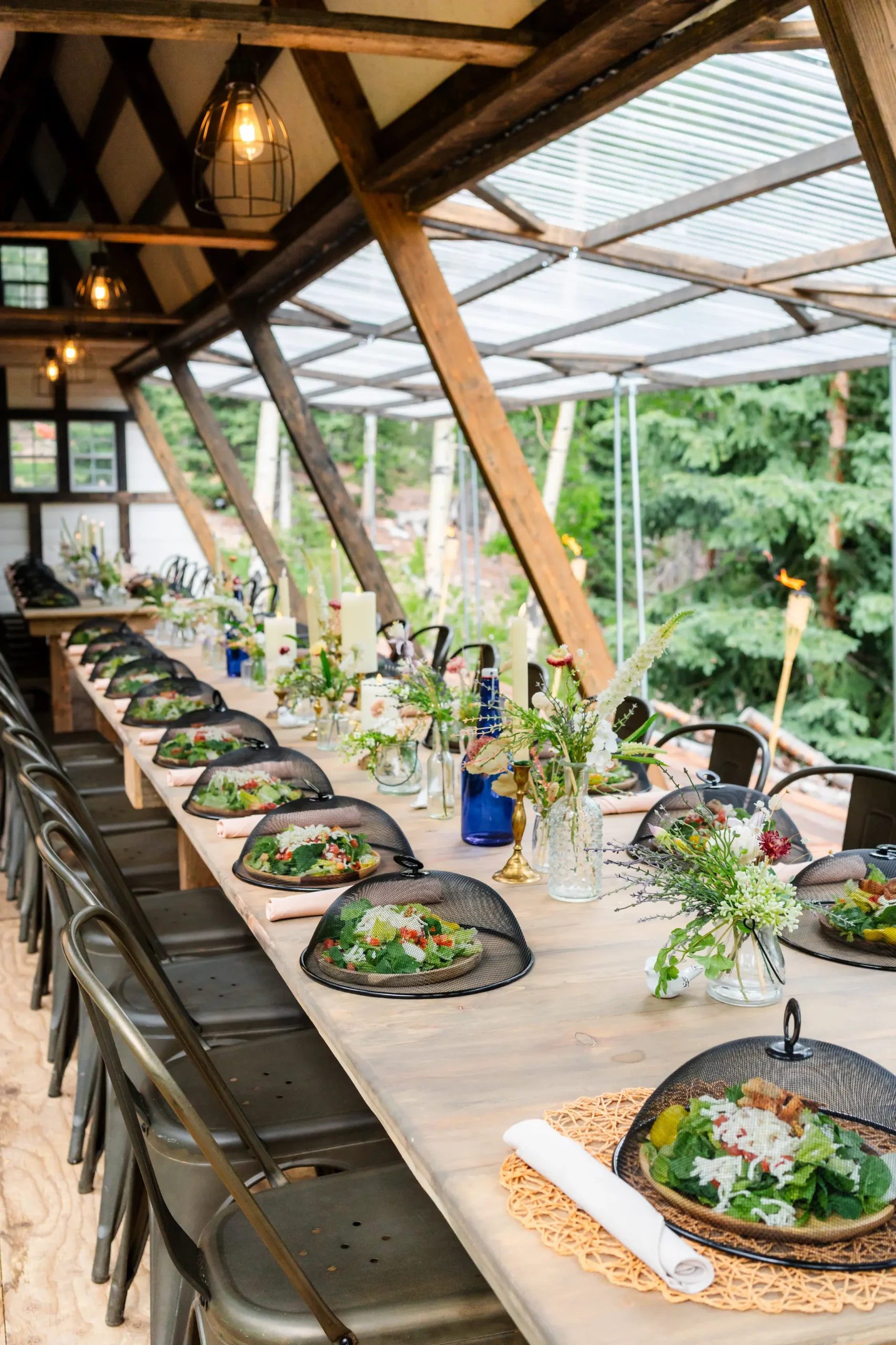Contact A beautifully set long wooden table for an outdoor wedding reception with floral centerpieces, place settings, and protective food covers.