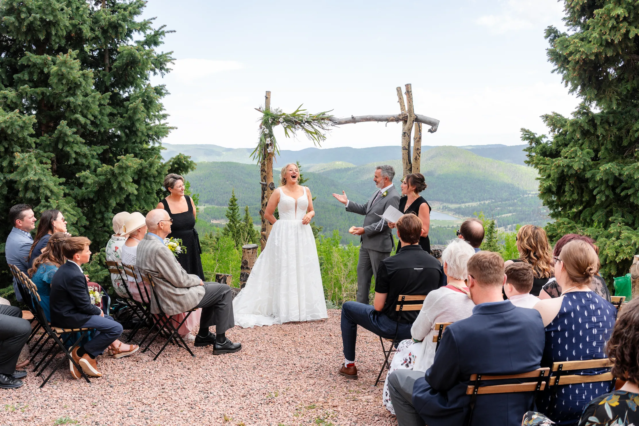 About Joyful bride laughing during an outdoor mountaintop wedding ceremony under a rustic wooden arbor, with guests seated in rows and a scenic mountain valley view in the background.